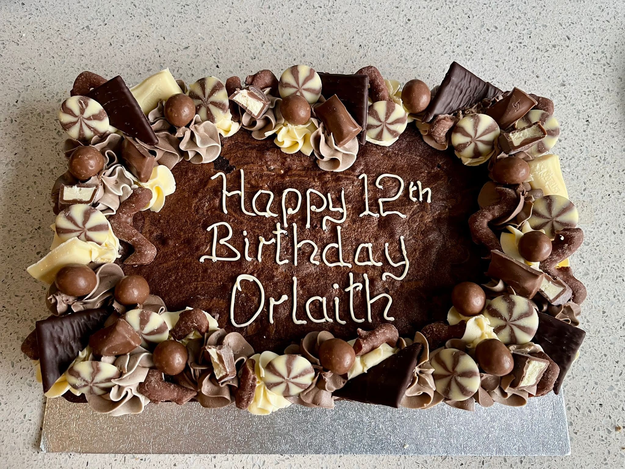 A rectangular chocolate brownie decorated with white icing spelling 'Happy 12th Birthday Orlaith', surrounded by swirls of light and dark chocolate frosting and a variety of chocolates including milk chocolate balls, white chocolate chunks, dark chocolate pieces, and peppermint creams.