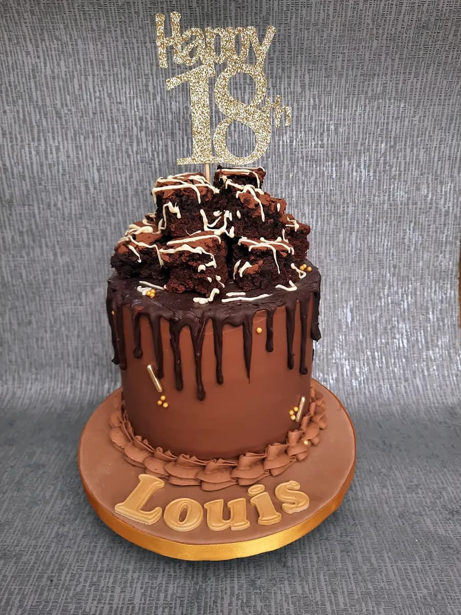A rich chocolate 18th birthday cake with a chocolate drip, topped with a stack of brownies drizzled with white icing, a gold glitter 'Happy 18th' topper, and gold 'Louis' lettering on the cake board.