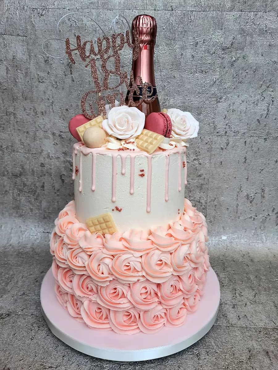 A two-tier 18th birthday cake featuring a white top tier with pink drip icing and sprinkles, and a pink bottom tier decorated with buttercream roses. The cake is adorned with a rose gold champagne bottle, a 'Happy 18th Birthday' topper, white sugar roses, pink macarons, and white chocolate bars.