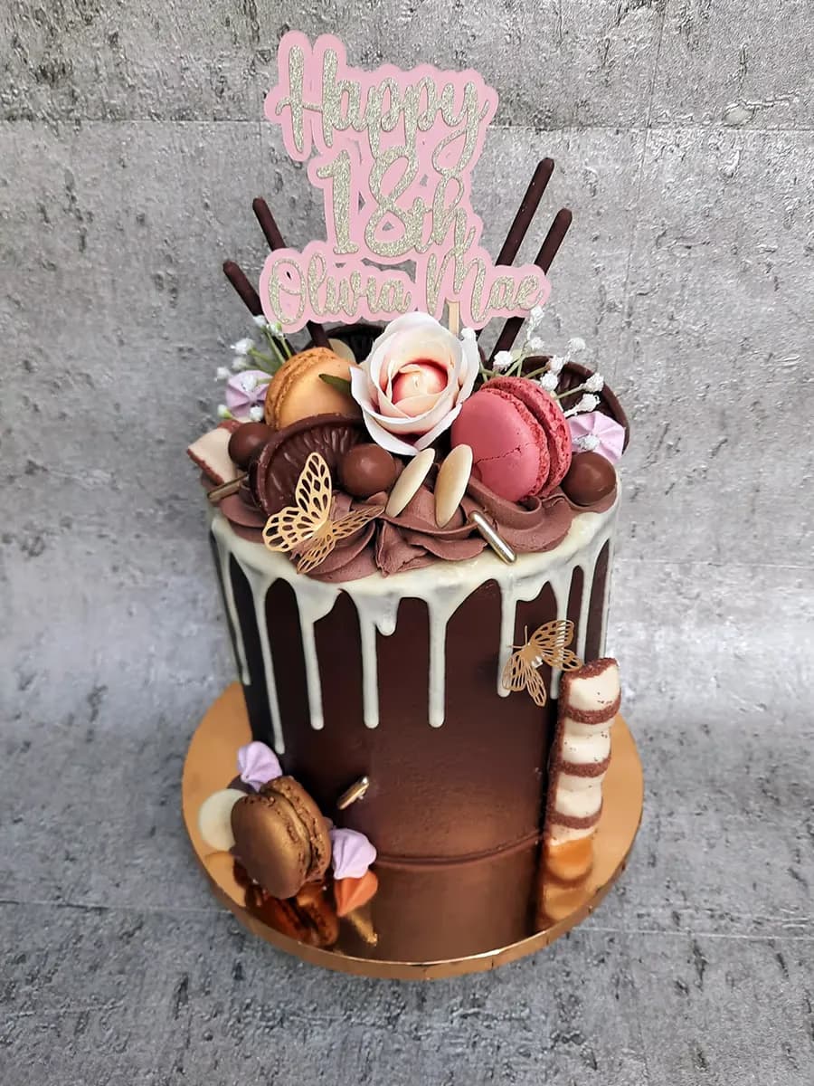 A dark chocolate drip cake for an 18th birthday, topped with a pink glitter 'Happy 18th Olivia Mae' topper, macarons, a white rose, chocolate decorations, and a Kinder Bueno bar on the side.