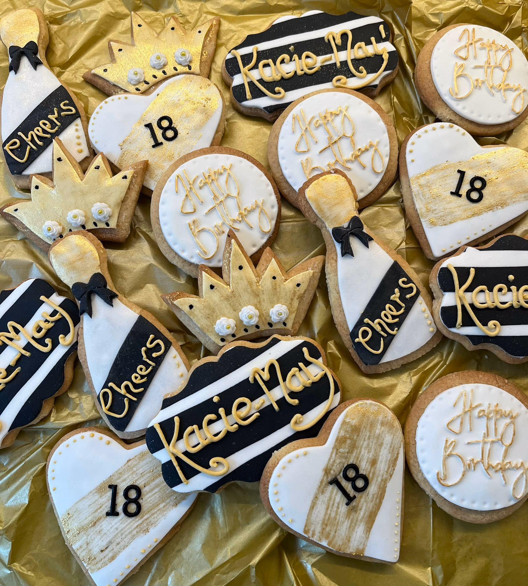 A close-up shot of a variety of black, white, and gold decorated sugar cookies for an 18th birthday, including champagne bottles, crowns, heart-shaped cookies with '18', round 'Happy Birthday' cookies, and striped cookies with 'Kacie May' in gold script, all arranged on a gold foil background.