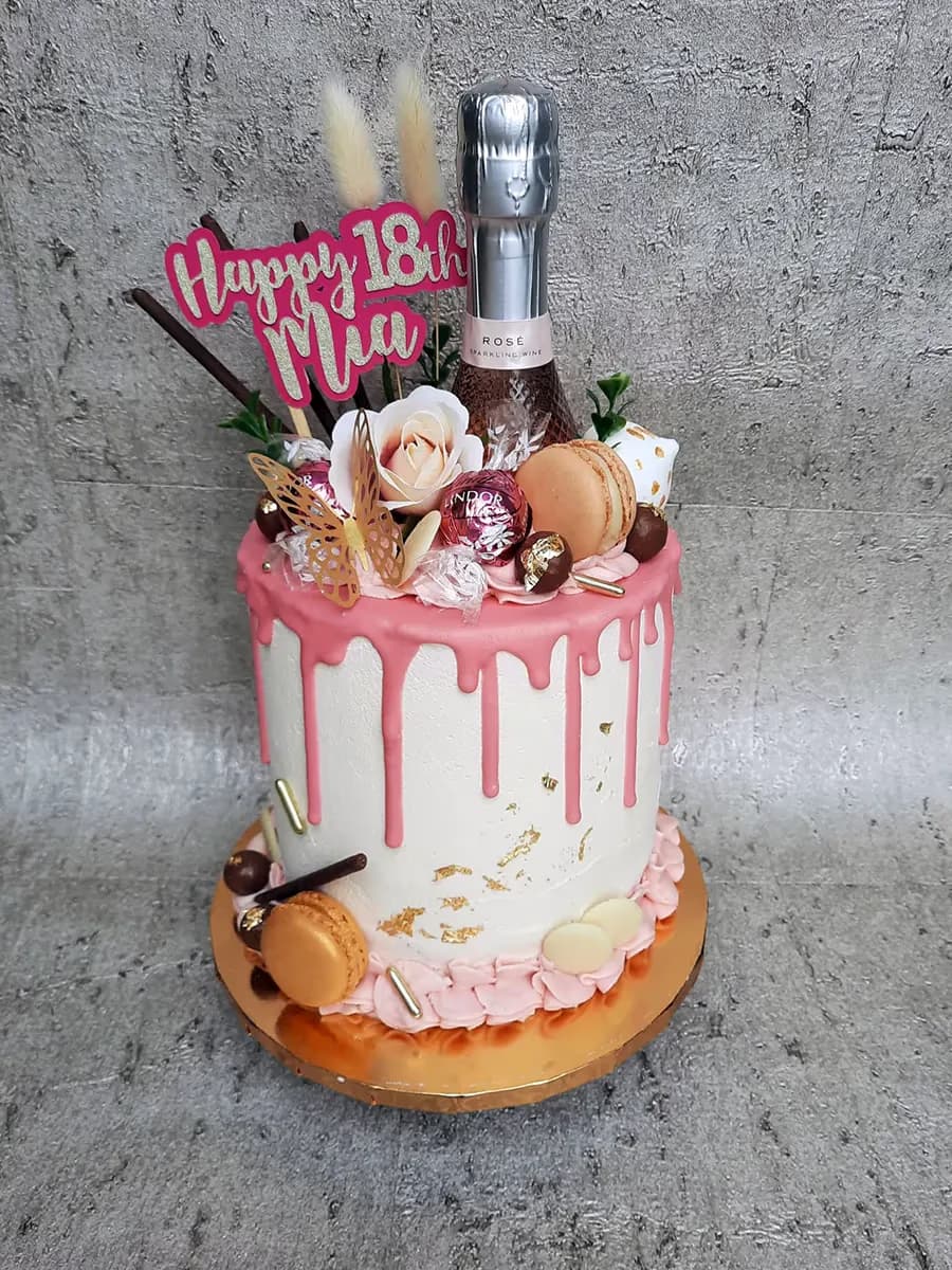 A white birthday drip cake with pink icing, decorated with a 'Happy 18th Mia' topper, a mini bottle of rosé sparkling wine, macarons, chocolates, a gold butterfly, white and pink artificial flowers, and gold flakes on the cake.