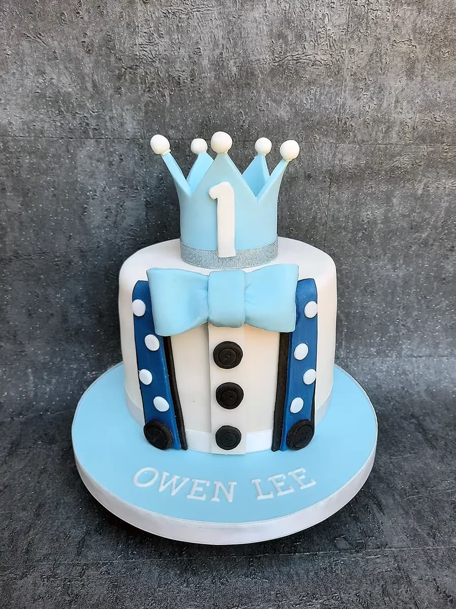 A white single-tier boy's first birthday cake decorated with light blue suspenders, a light blue bow tie, and black buttons, topped with a light blue crown featuring the number one. The light blue cake board has 'Owen Lee' written on it.