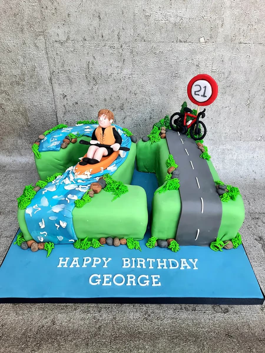 A vibrant 21st birthday cake for George, shaped like the number 21, featuring a person kayaking on a blue river on the '2' and a red bicycle with a '21' speed limit sign on a grey road on the '1', all set on a blue cake board with the text 'HAPPY BIRTHDAY GEORGE'.