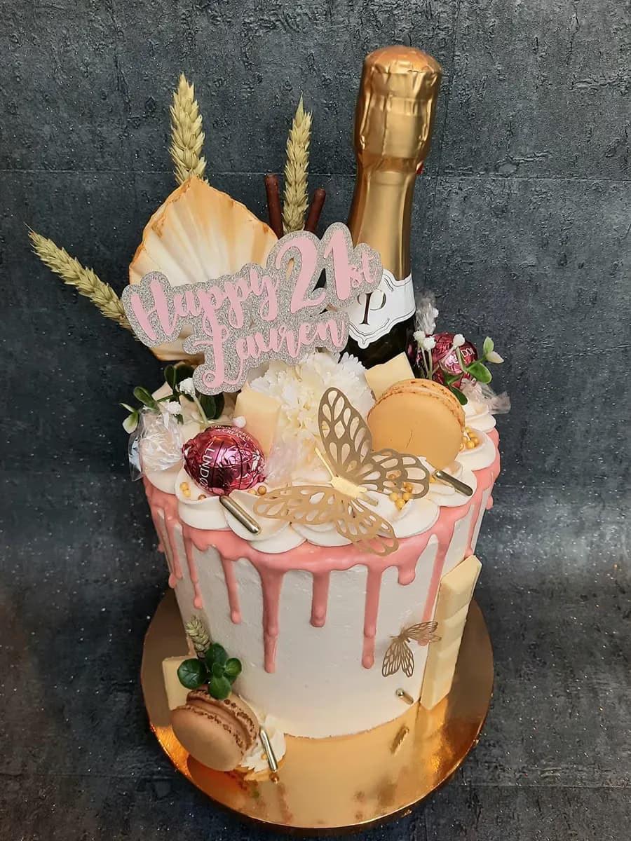 A single-tier 21st birthday cake with white frosting, pink drip icing, and a 'Happy 21st Lauren' topper. It is decorated with a miniature gold champagne bottle, gold butterflies, macarons, white chocolate pieces, and dried wheat stalks, all on a gold cake board.