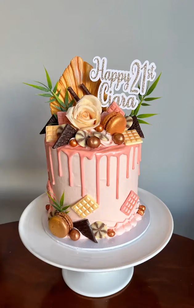 A light pink 21st birthday drip cake, decorated with a 'Happy 21st Ciara' topper, a peach rose, gold macarons, various chocolates, and green foliage, displayed on a white cake stand.