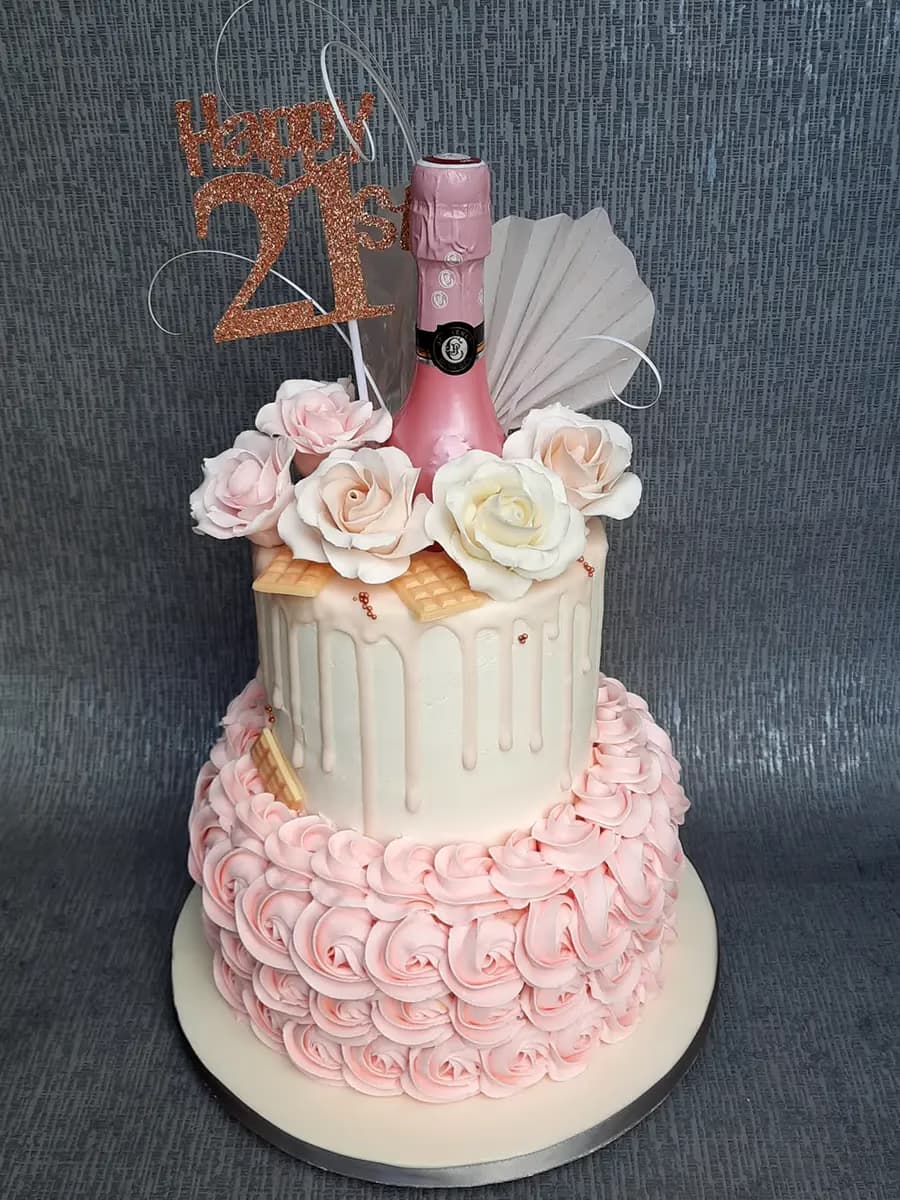 A two-tier 21st birthday cake with a pink rosette bottom tier and a white drip top tier, topped with a mini pink champagne bottle, pink and white sugar roses, and a glittery 'Happy 21st' topper.