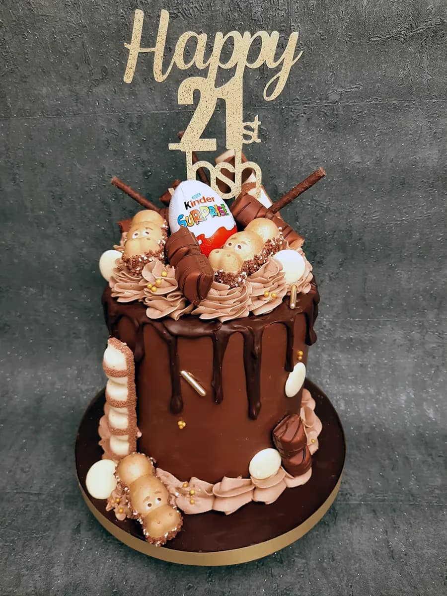 A rich chocolate drip cake for a 21st birthday, adorned with a gold 'Happy 21st' topper, a Kinder Surprise egg, Kinder Happy Hippo chocolates, and various other Kinder treats.