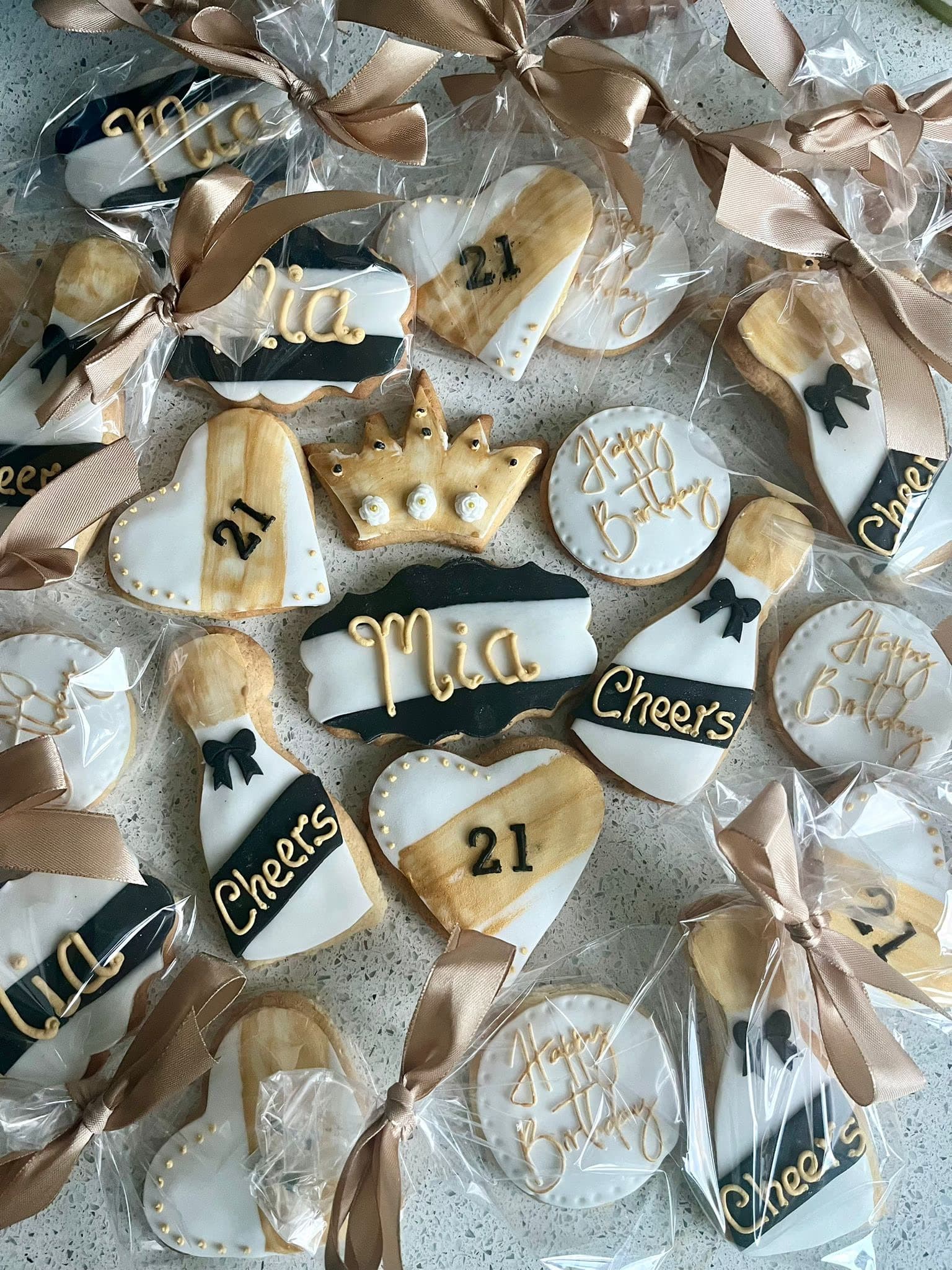 A collection of individually wrapped and ribbon-tied 21st birthday themed cookies in black, white, and gold, featuring designs like 'Mia', '21', 'Cheers', 'Happy Birthday', and a crown, on various shapes including hearts, champagne bottles, and plaques.