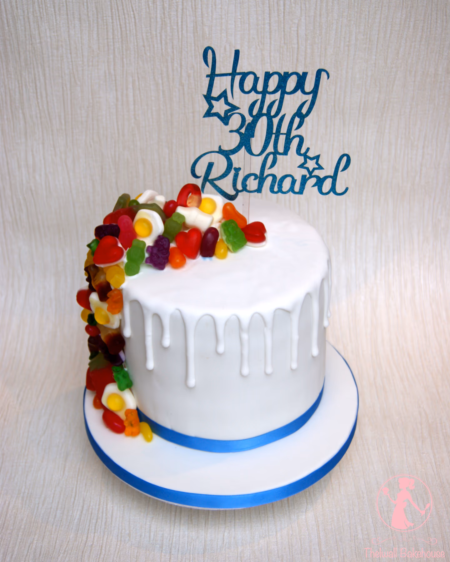 A white round birthday cake with white drip icing, decorated with a cascade of colorful gummy candies, and topped with a blue glittery 'Happy 30th Richard' sign, sitting on a white board with a blue ribbon.