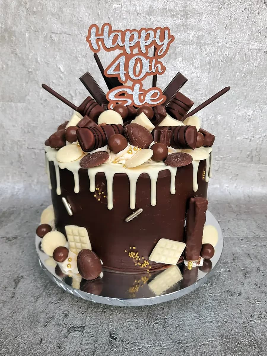 A rich dark chocolate drip cake for a 40th birthday, adorned with a 'Happy 40th Ste' topper and generously decorated with an assortment of milk and white chocolates, including Kinder Bueno, chocolate balls, and white chocolate pieces, finished with gold and silver sprinkles.