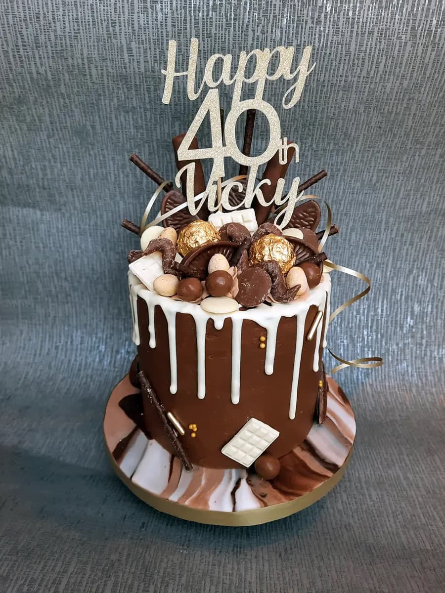 A tall chocolate drip cake with white chocolate drips, topped with an assortment of chocolates, gold foil candies, chocolate sticks, and a gold glitter "Happy 40th Vicky" cake topper. The cake sits on a marble-effect base with a gold rim.