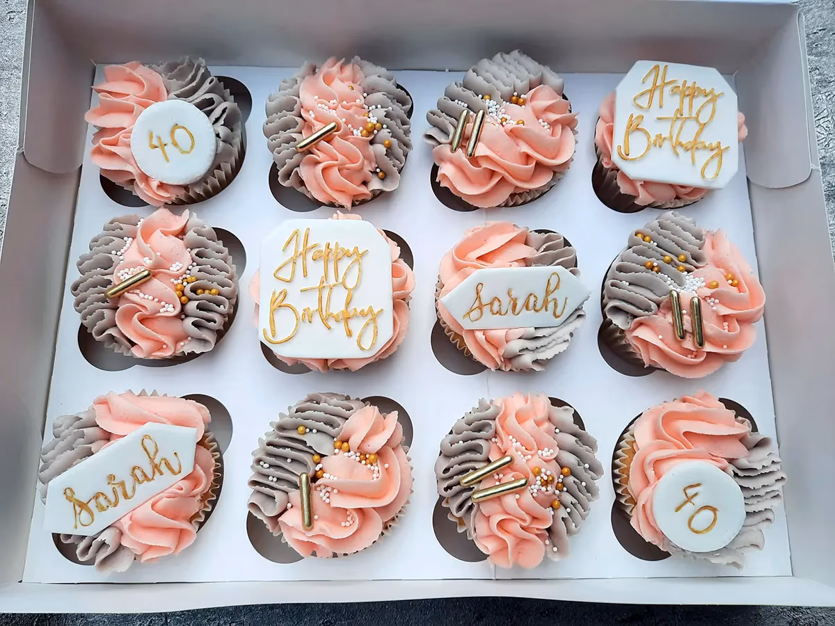 A dozen cupcakes in a white box, decorated with alternating pink and grey frosting swirls, gold sprinkles, and gold bar sprinkles. Some cupcakes feature white fondant toppers with gold text, including 'Happy Birthday', 'Sarah', and '40'.