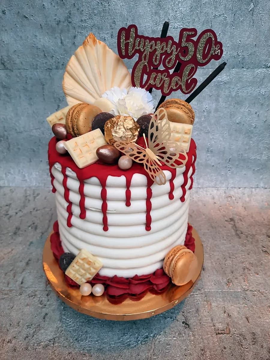 A round white ribbed 50th birthday cake for Carol, featuring a vibrant red drip glaze. It is elaborately decorated with gold macarons, various chocolates including a gold-wrapped truffle, white chocolate bar pieces, a gold cut-out butterfly, small white flowers, a cream-colored fan decoration, and a 'Happy 50th Carol' glitter cake topper. The cake sits on a gold cake board with red ruffled frosting at the base.