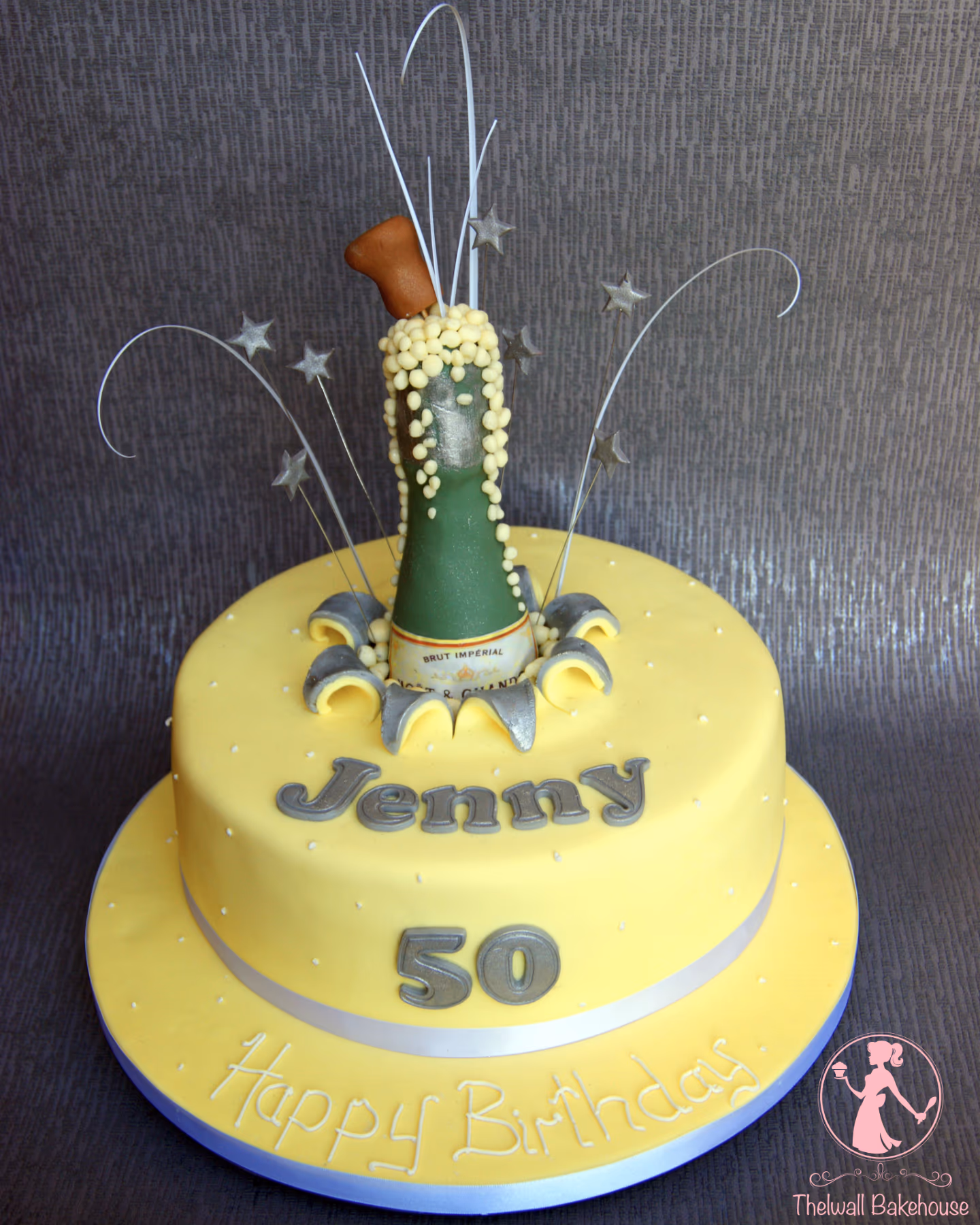 A vibrant yellow two-tier 50th birthday cake featuring a champagne bottle topper exploding with white sugar pearls and silver decorative stars. The cake is inscribed with 'Jenny' and '50' in silver lettering, and 'Happy Birthday' in white script on the cake board.