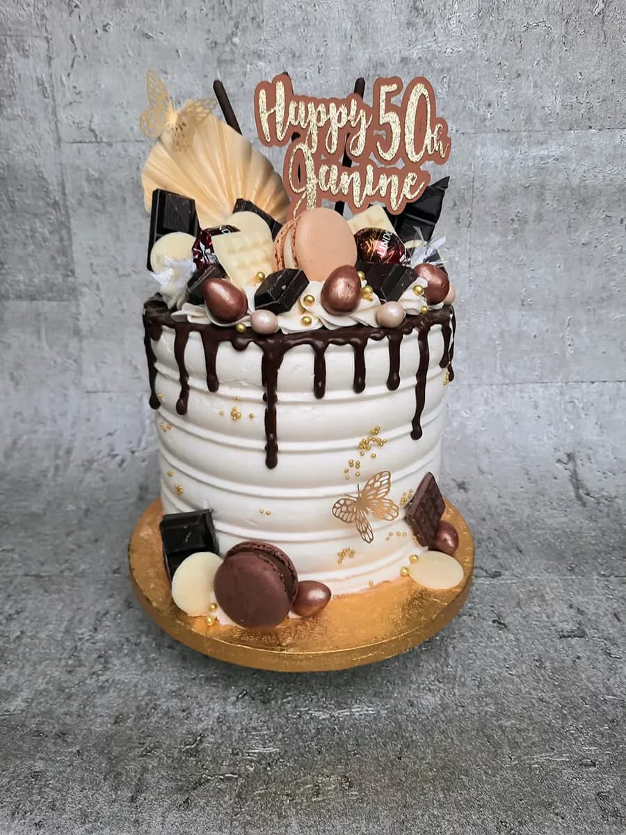 A single-tier white ridged birthday cake with a dark chocolate drip, lavishly decorated on top with various chocolates, light and dark macarons, gold edible balls, a large gold butterfly, and a 'Happy 50th Janine' cake topper. The base also features smaller chocolates, a dark macaron, and a gold butterfly on a gold cake board.