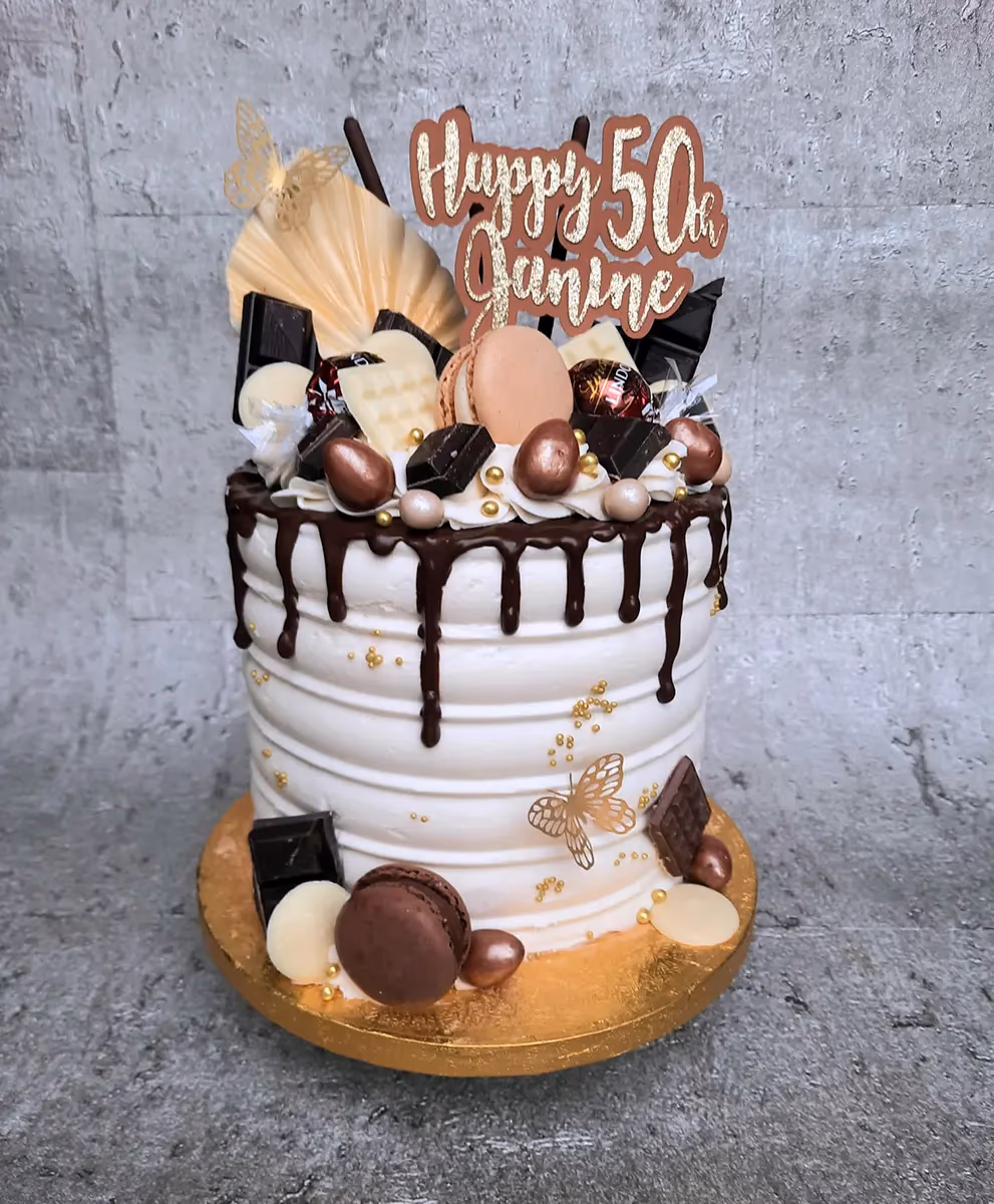 A white, ribbed birthday cake with a chocolate drip, decorated with various chocolates, macarons, gold sprinkles, gold butterflies, and a 'Happy 50th Janine' cake topper.