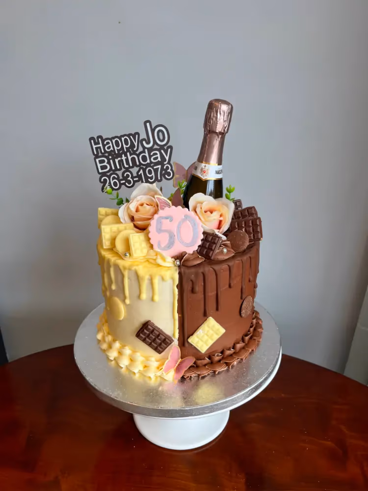 A two-tone 50th birthday cake with a vertical split design. One half features white chocolate drip and white chocolate pieces, while the other half has dark chocolate drip and dark chocolate pieces. The cake is decorated with a miniature rose champagne bottle, peach roses, a pink '50' disc, and a 'Happy Jo Birthday 26-3-1973' cake topper.