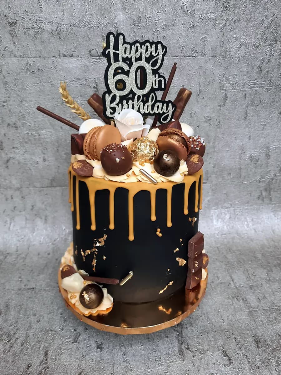A striking single-tier 60th birthday cake with black fondant, gold drips, and gold leaf accents, topped with a 'Happy 60th Birthday' sign, brown macarons, chocolate truffles, white roses, and various chocolate decorations.