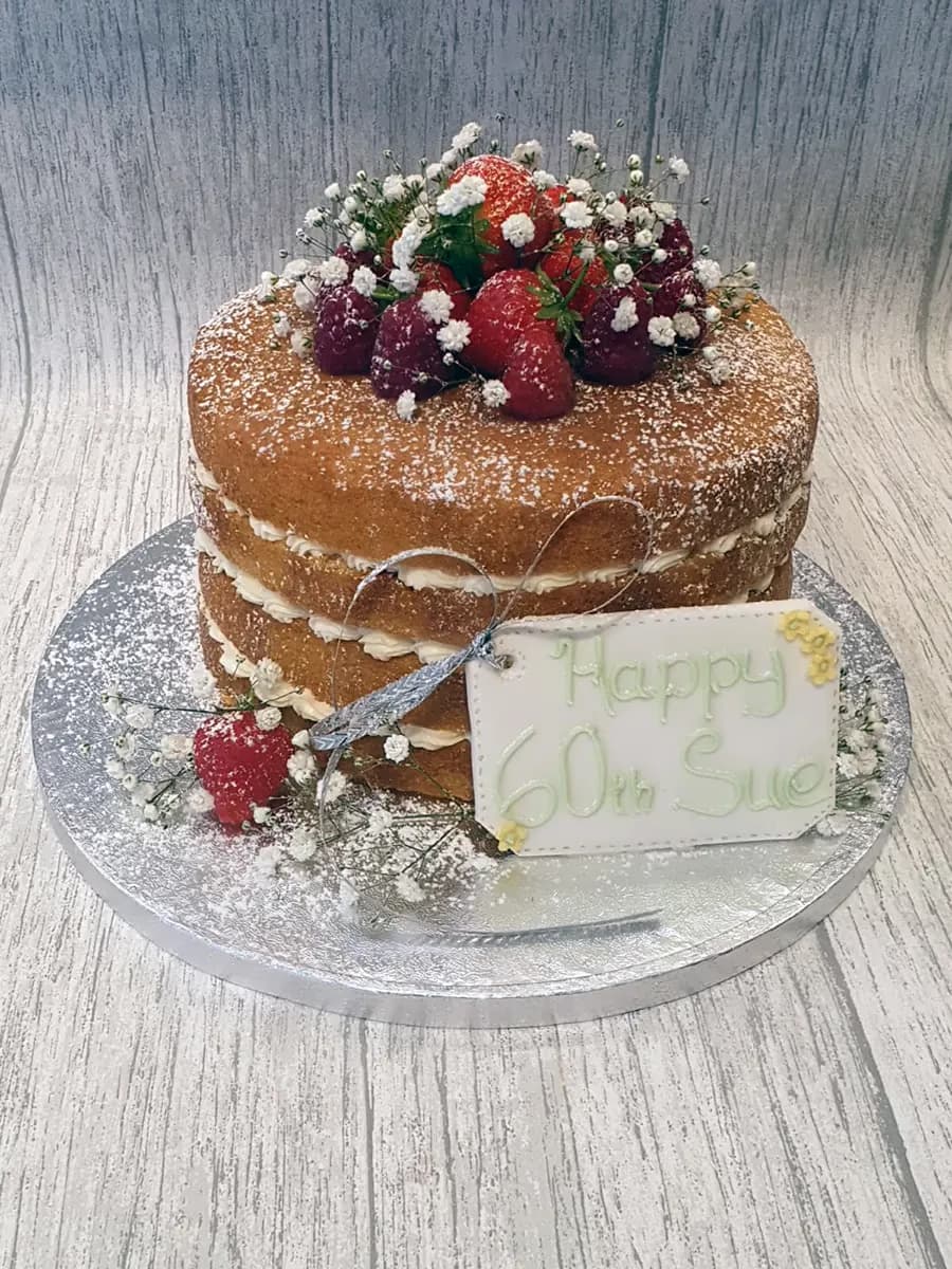 A two-tier naked sponge cake for a 60th birthday, dusted with powdered sugar, and decorated with fresh strawberries, raspberries, white baby's breath flowers, and a 'Happy 60th Sue' message tag.