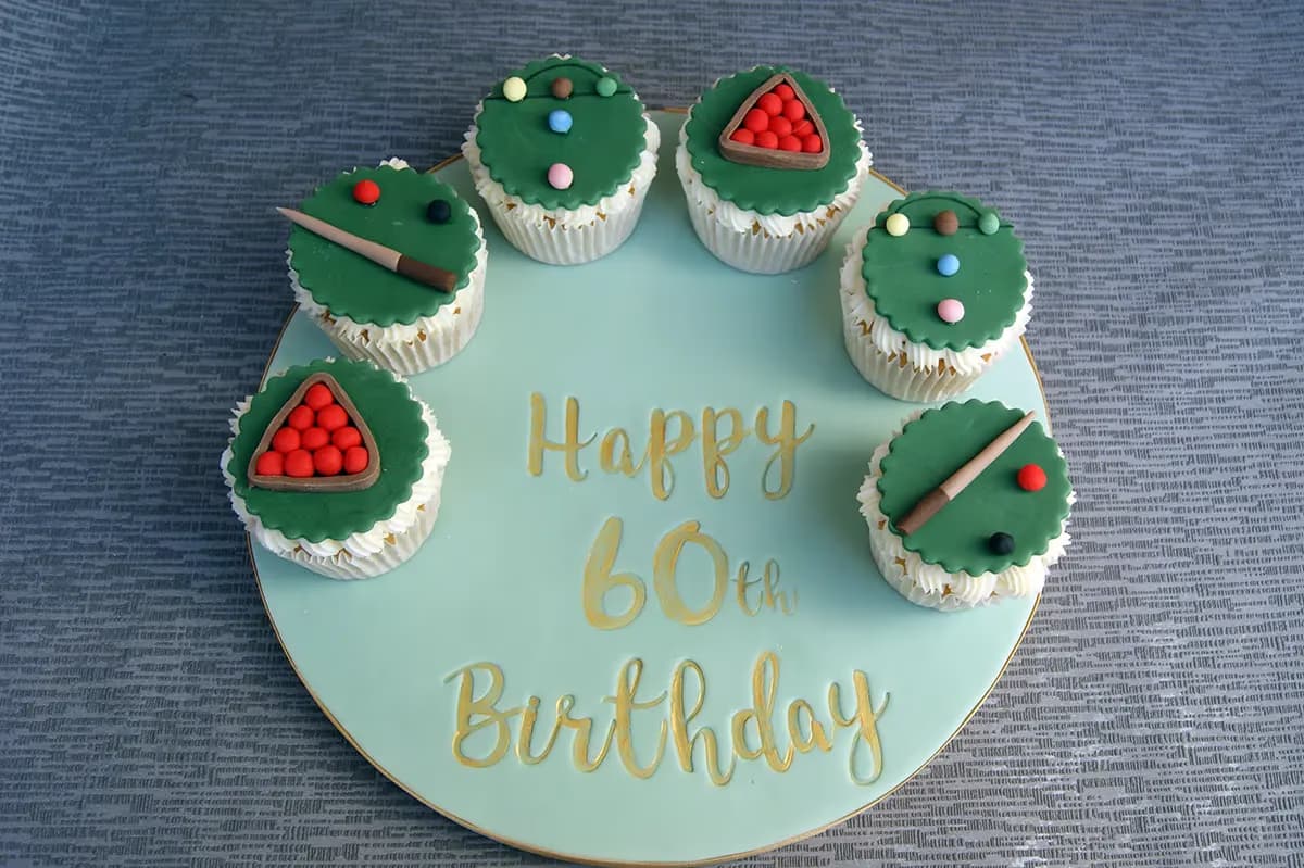 A light green round cake board with 'Happy 60th Birthday' in gold lettering, surrounded by six snooker or pool-themed cupcakes with green fondant tops, some featuring a red ball rack, others a cue and balls.