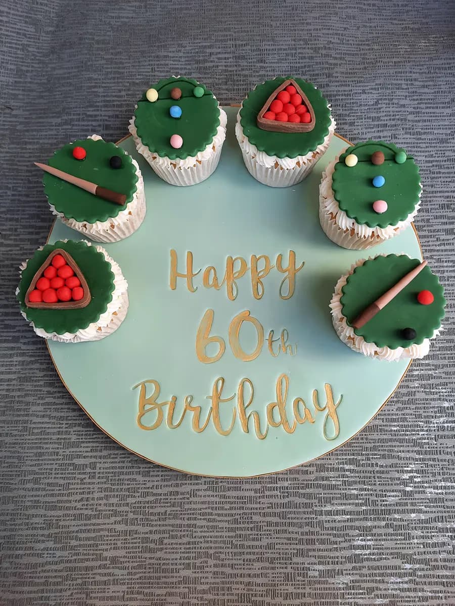 Six snooker or pool-themed cupcakes arranged on a round light green cake board with 'Happy 60th Birthday' written in gold. The cupcakes are decorated with green fondant, miniature cues, colored balls, and racks of red balls.