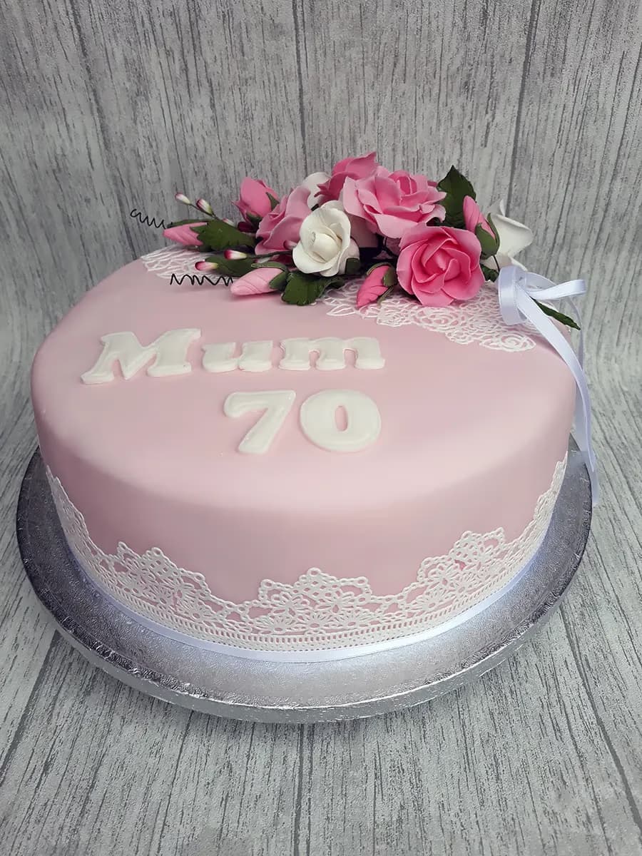 A round pink 70th birthday cake for 'Mum', decorated with white sugar letters, a bouquet of pink and white sugar roses, and white edible lace around the base.