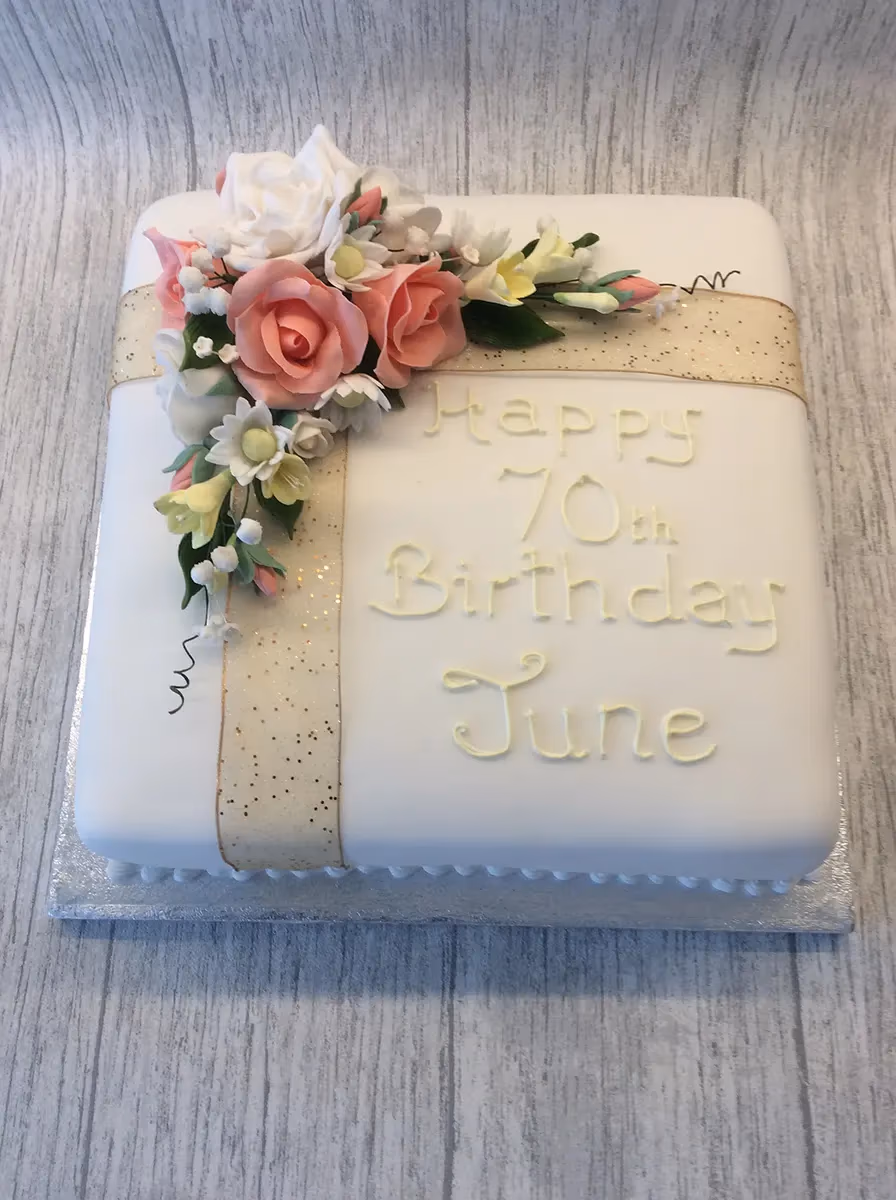 A square white fondant 70th birthday cake, inscribed with 'Happy 70th Birthday June', decorated with an arrangement of pink and white sugar roses and small yellow flowers, and a glittery gold ribbon.