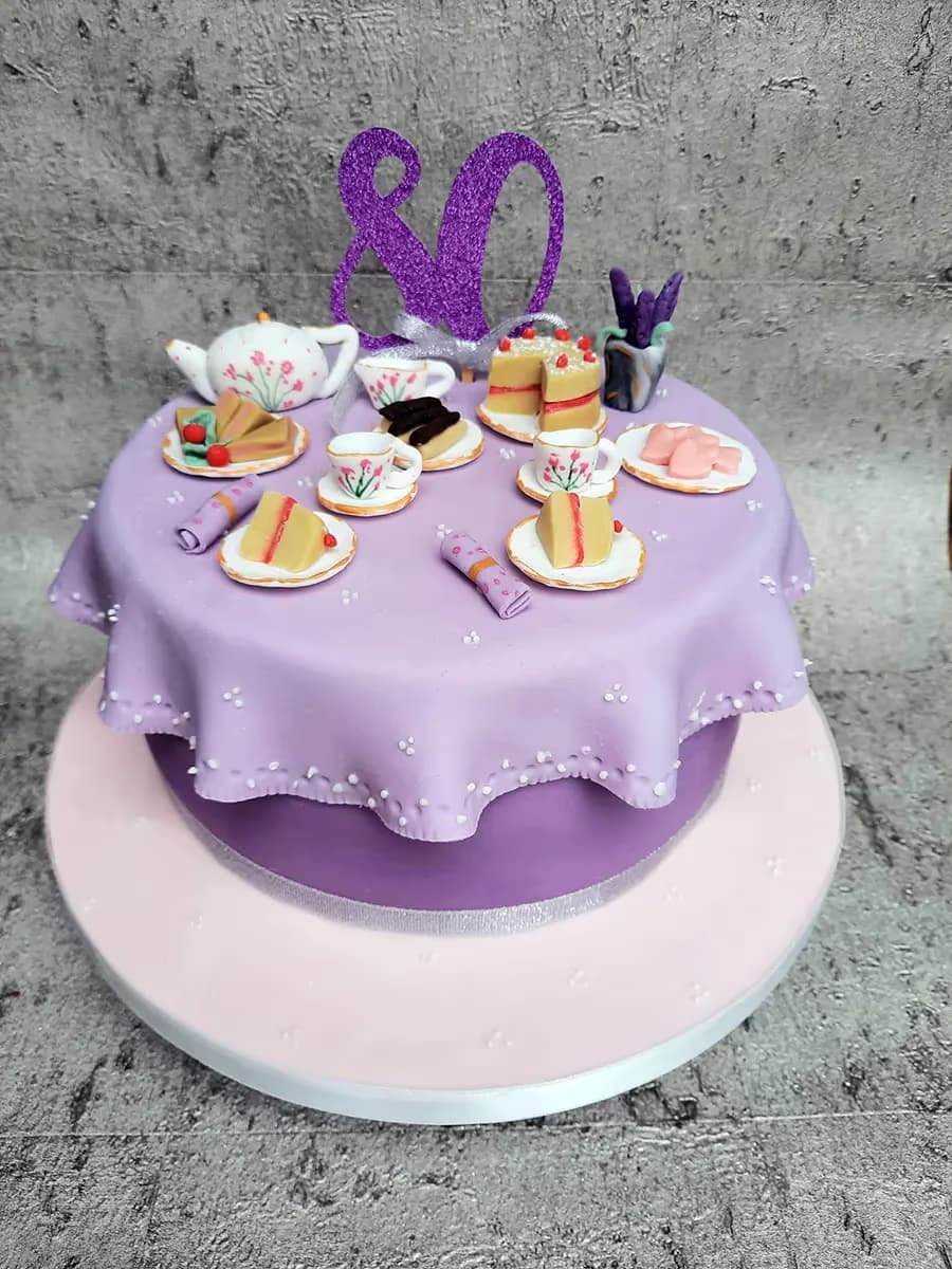 A two-tier 80th birthday cake, with the top tier decorated as a miniature tea party table featuring a purple tablecloth, a glittery '80' topper, a teapot, teacups, cake slices, and pastries. The cake sits on a light pink cake board.