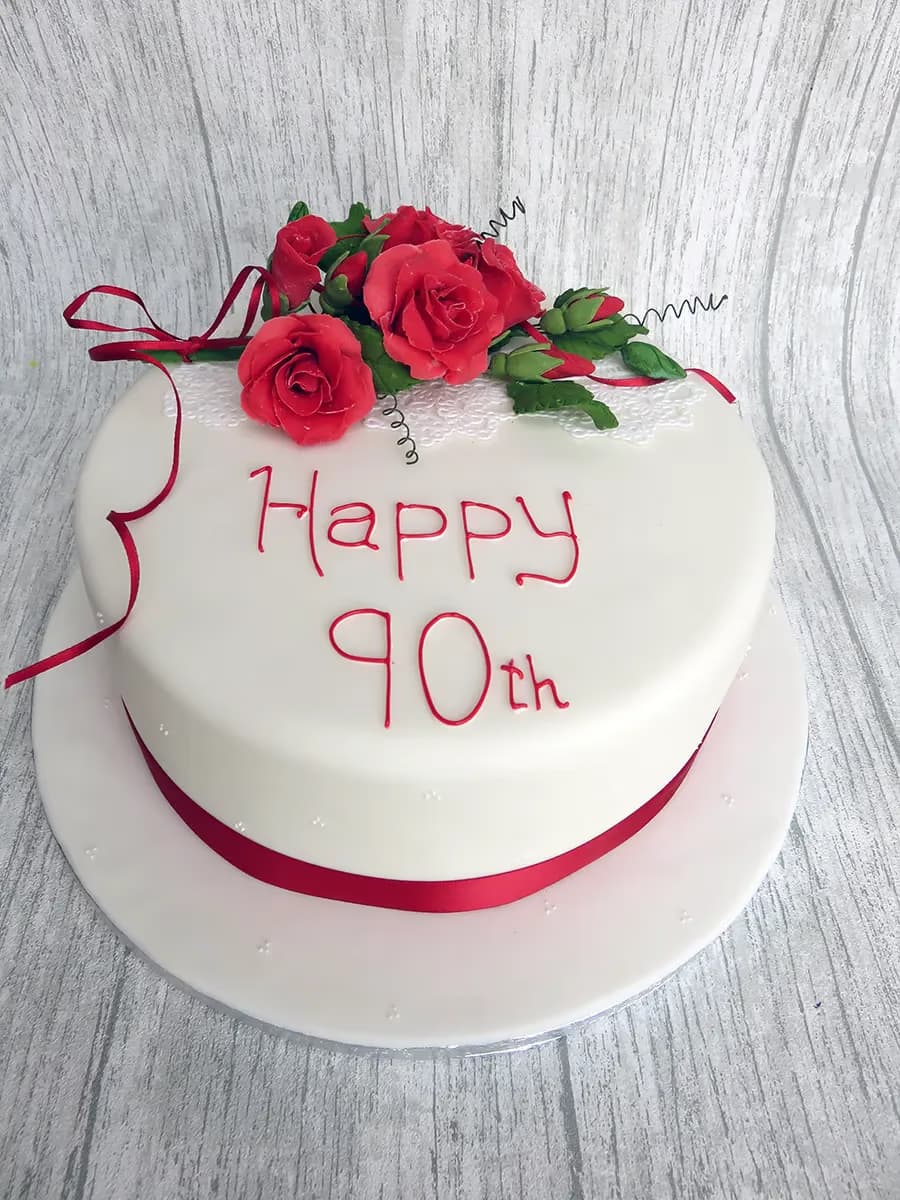 A round white fondant cake for a 90th birthday, decorated with a bouquet of red sugar roses, a red ribbon around the base, and 'Happy 90th' written in red icing.