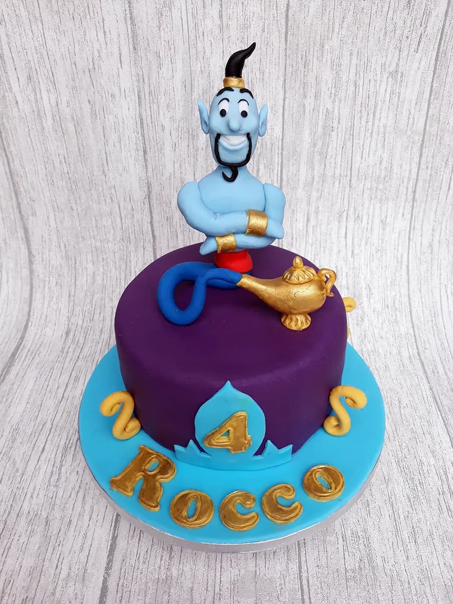 An Aladdin-themed birthday cake featuring a blue Genie figure with crossed arms, a golden magic lamp, a number 4, and the name 'Rocco' on a purple cake with a light blue base.