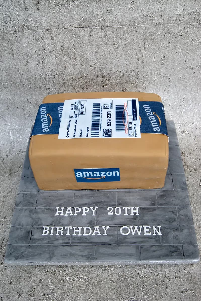 A novelty birthday cake shaped like a brown Amazon package with blue Amazon tape and a shipping label, sitting on a grey tiled base inscribed with 'Happy 20th Birthday Owen'.