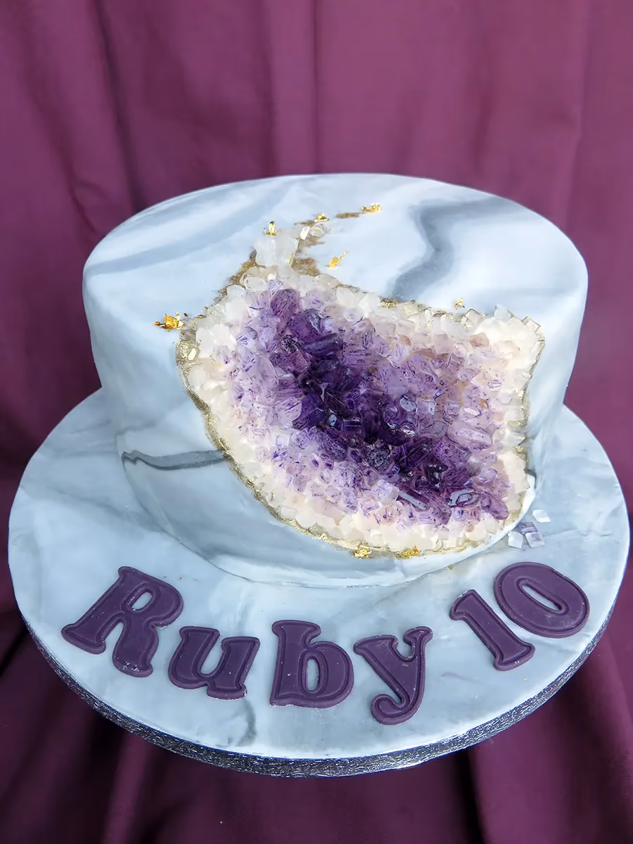 A single-tier cake decorated with a grey and white marble effect, featuring a large amethyst geode design on top with deep purple and lighter crystal formations, outlined with gold and gold leaf details. The cake board has 'Ruby 10' written in purple letters.