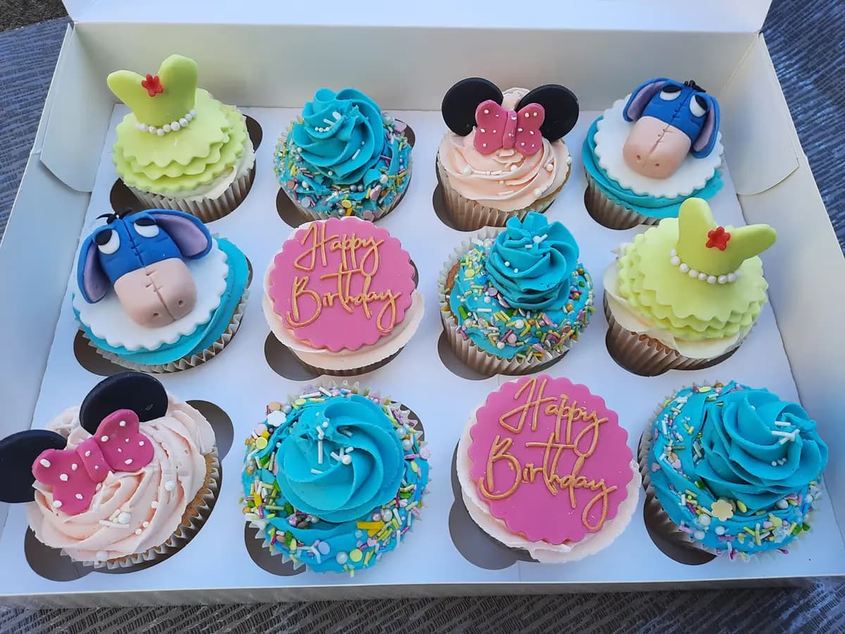 A dozen assorted themed cupcakes in a white box, featuring designs of Minnie Mouse, Eeyore, green princess dresses, blue frosting with colorful sprinkles, and pink 'Happy Birthday' toppers.