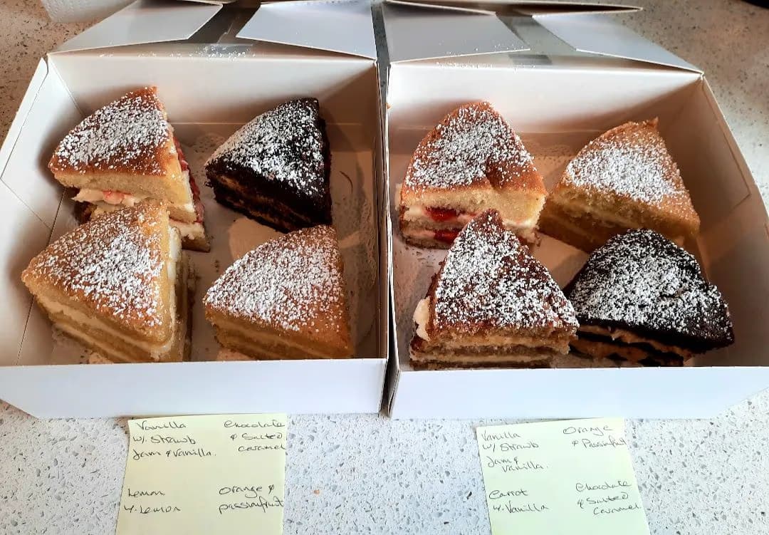 Eight triangular mini cake slices, each dusted with powdered sugar, presented in two white boxes. The assorted flavors include vanilla with strawberry jam, chocolate with salted caramel, lemon, orange with passionfruit, and carrot with vanilla.