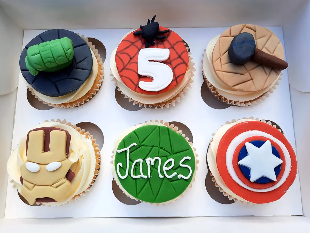 Six Avengers superhero themed cupcakes in a white box. The cupcakes feature decorations of Hulk's green fist, Spider-Man's web with the number 5 and a black spider, Thor's hammer, Iron Man's mask, Captain America's shield, and a green cupcake with 'James' written on it.
