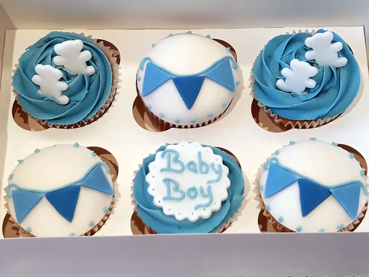 Six blue and white baby boy cupcakes in a box. Two have blue frosting with white teddy bear toppers, three have white fondant tops with blue bunting, and one has blue frosting with a white plaque saying 'Baby Boy'.
