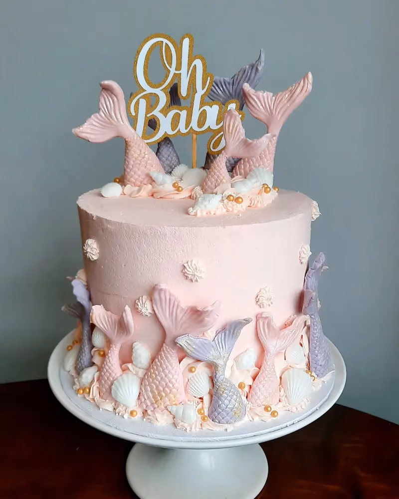 A pink single-tier baby shower cake decorated with light pink and lavender mermaid tails, white seashells, gold sprinkles, and a gold and white 'Oh Baby' cake topper.