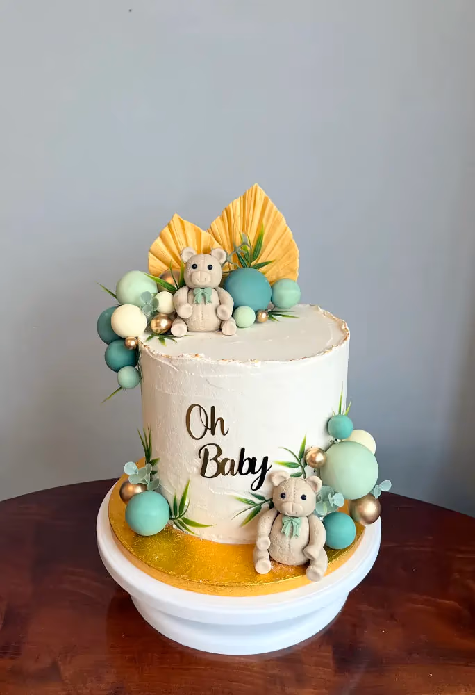 A single-tier white baby shower cake decorated with two teddy bears, green, white, and gold spheres, yellow fan-shaped decorations, and green foliage, with 'Oh Baby' written on the side.