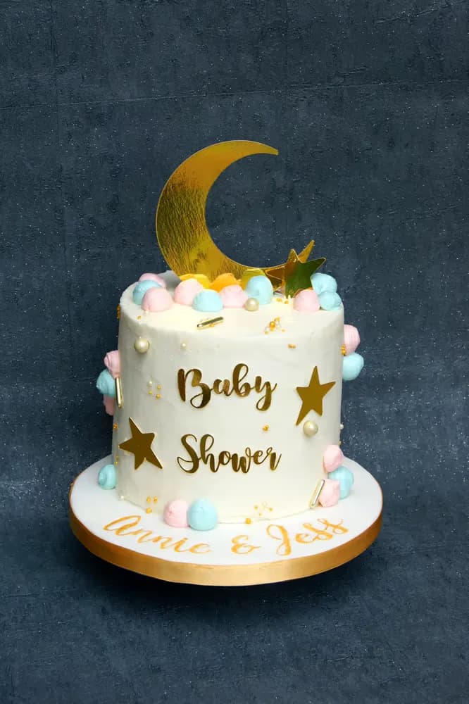 A white baby shower cake decorated with a gold moon and star topper, gold 'Baby Shower' lettering, gold star cutouts, and pastel pink, blue, and yellow meringue kisses, with 'Annie & Jess' written on the gold-edged base.