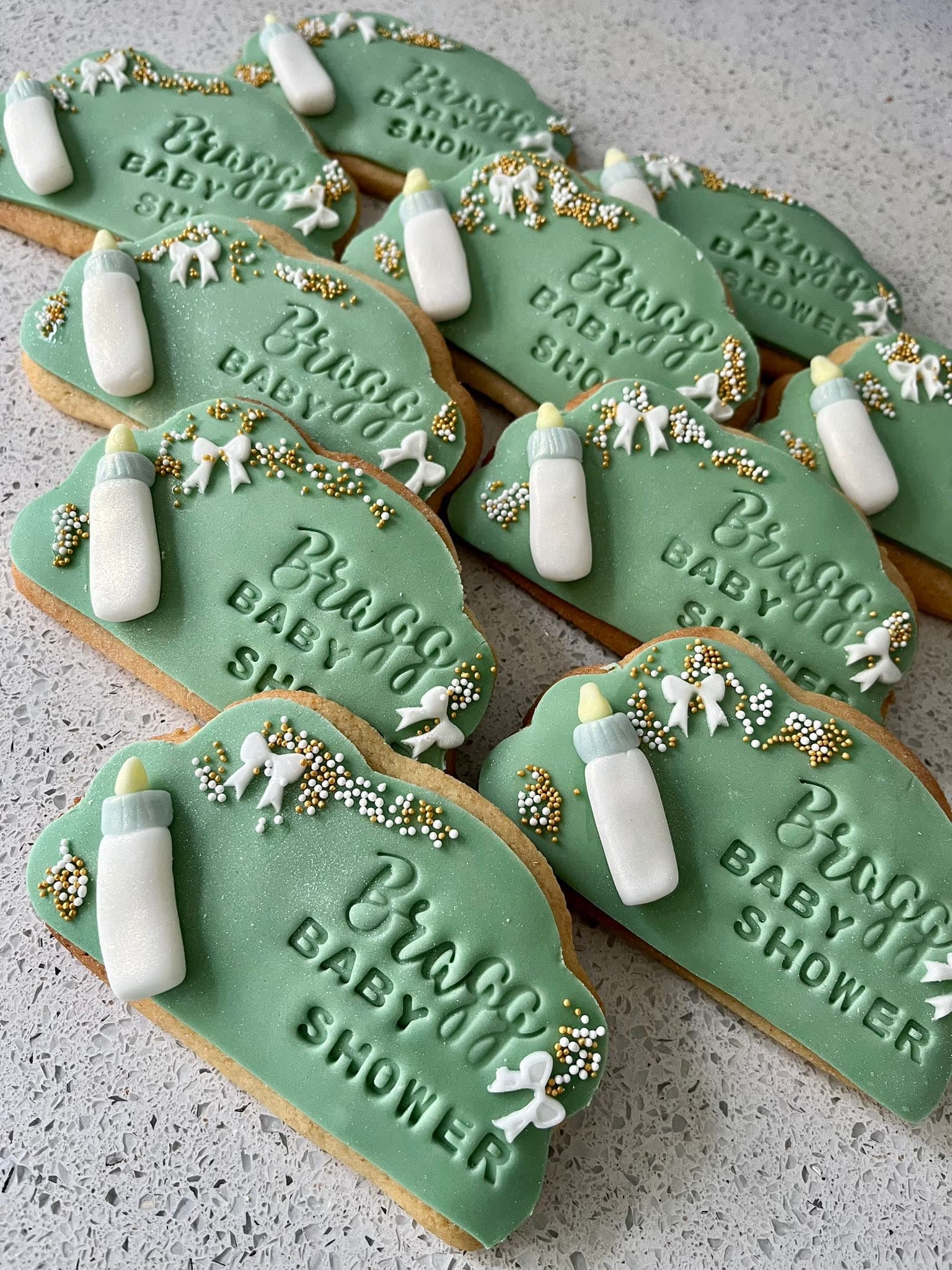 Close-up of multiple green plaque-shaped baby shower cookies, each featuring a white baby bottle, white fondant bows, gold and white sprinkles, and the embossed text 'Braggs BABY SHOWER'.