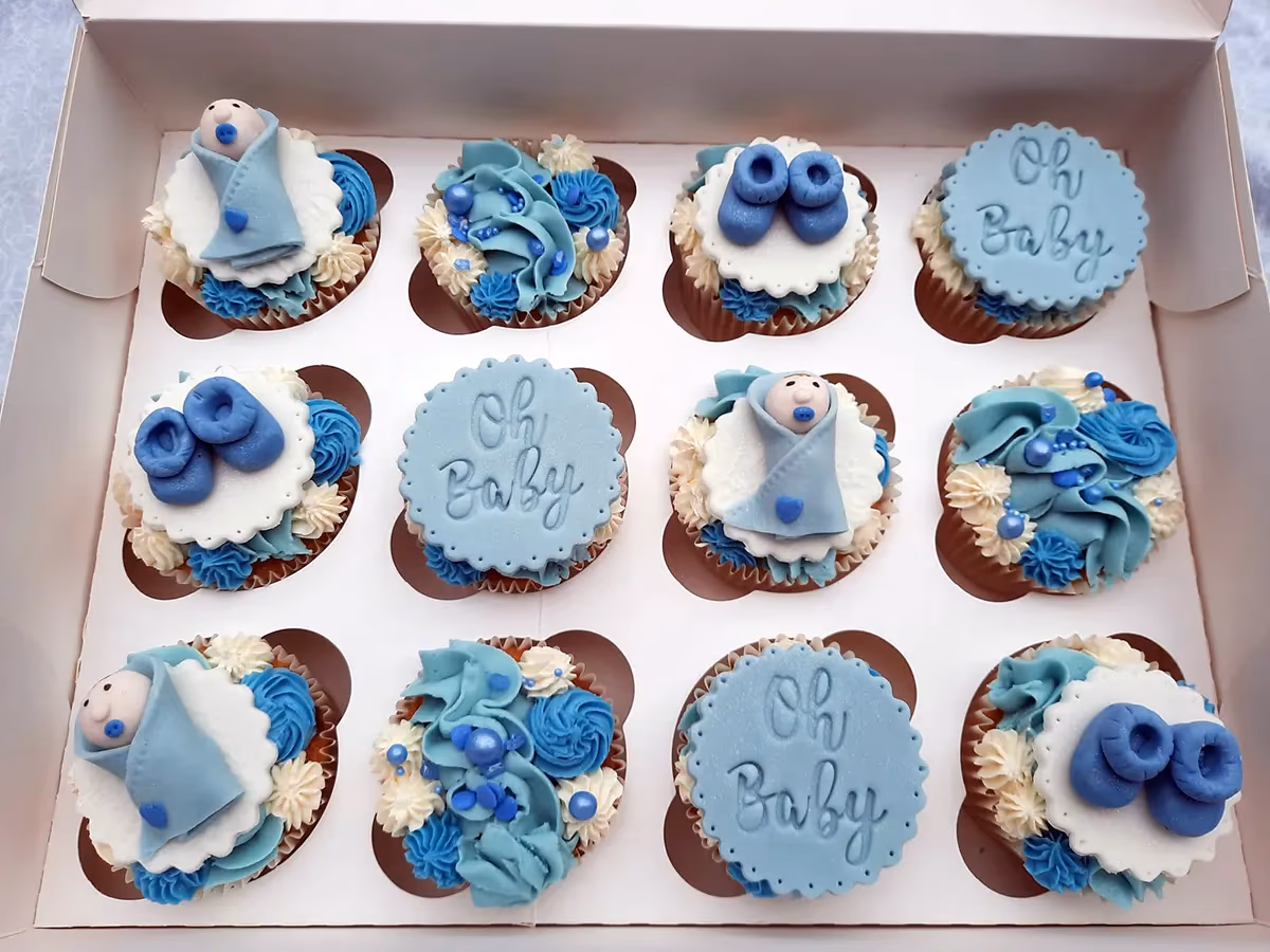 A box of twelve blue and white baby shower cupcakes with mixed designs, including baby figures, baby booties, 'Oh Baby' text, and decorative frosting swirls.