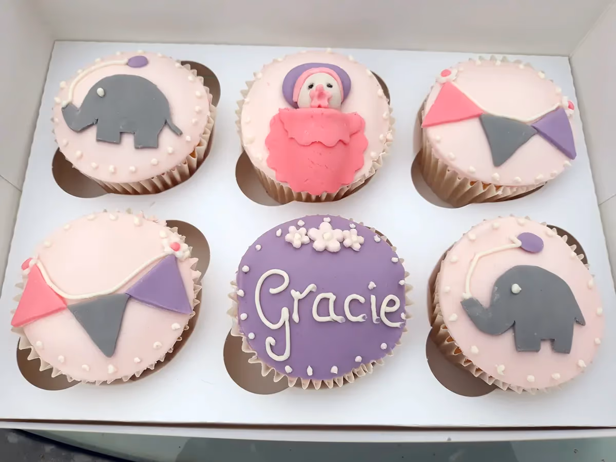 Six baby shower themed cupcakes arranged in a white box. Two cupcakes feature a grey elephant with a purple balloon, two have pink, grey, and purple bunting flags, one has a pink baby figure in a swaddle, and one purple cupcake is decorated with 'Gracie' written in white script and small white flowers. The pink-topped cupcakes have white polka dot borders.