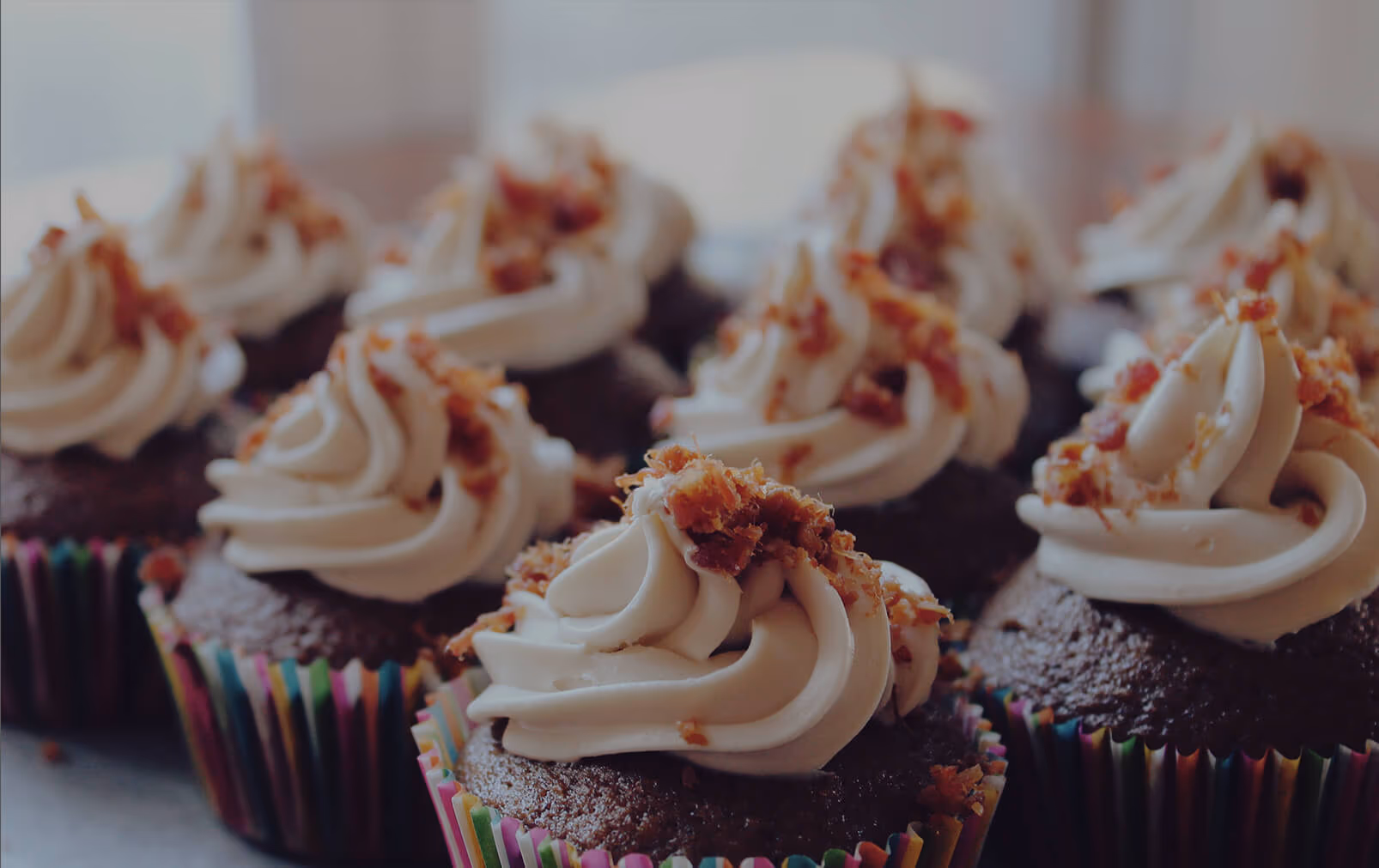 Several chocolate cupcakes topped with white swirled frosting and crumbled bacon, presented in colorful striped wrappers.