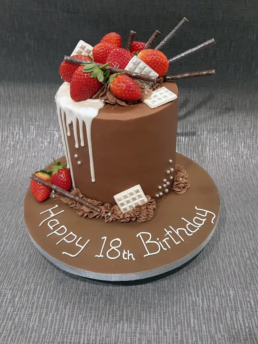 A single-tier chocolate birthday cake decorated with fresh strawberries, chocolate sticks, white chocolate pieces, white drip icing, and silver pearls, with 'Happy 18th Birthday' written on the cake board.