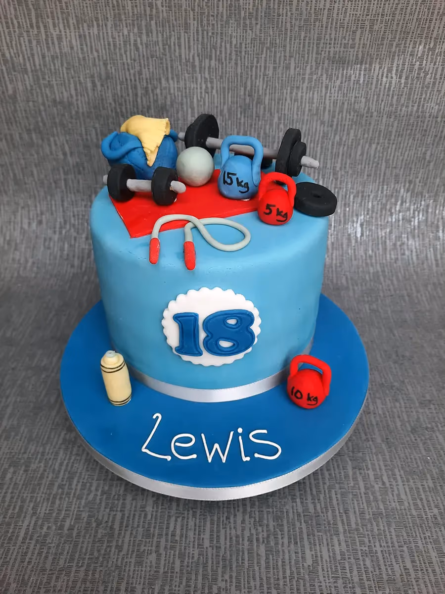 A light blue single-tier birthday cake with a gym and fitness theme, decorated with miniature barbells, kettlebells, a skipping rope, and a water bottle. The cake has '18' on the side and 'Lewis' written on the blue cake board.