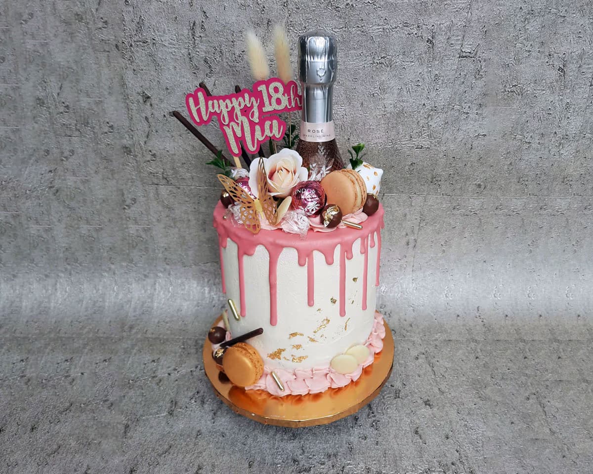 A single-tier birthday cake with white frosting and pink drip icing, decorated with a 'Happy 18th Mia' cake topper, a miniature champagne bottle, macarons, chocolates, artificial flowers, gold butterfly accents, and gold flakes.
