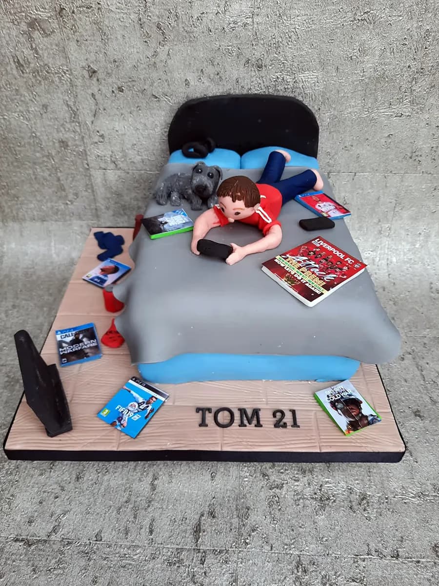 A 21st birthday cake featuring a fondant figure of a man lying on a bed, playing a video game with a controller, next to a grey dog. The bed is surrounded by miniature video game cases and a Liverpool FC book. The cake base reads 'TOM 21'.