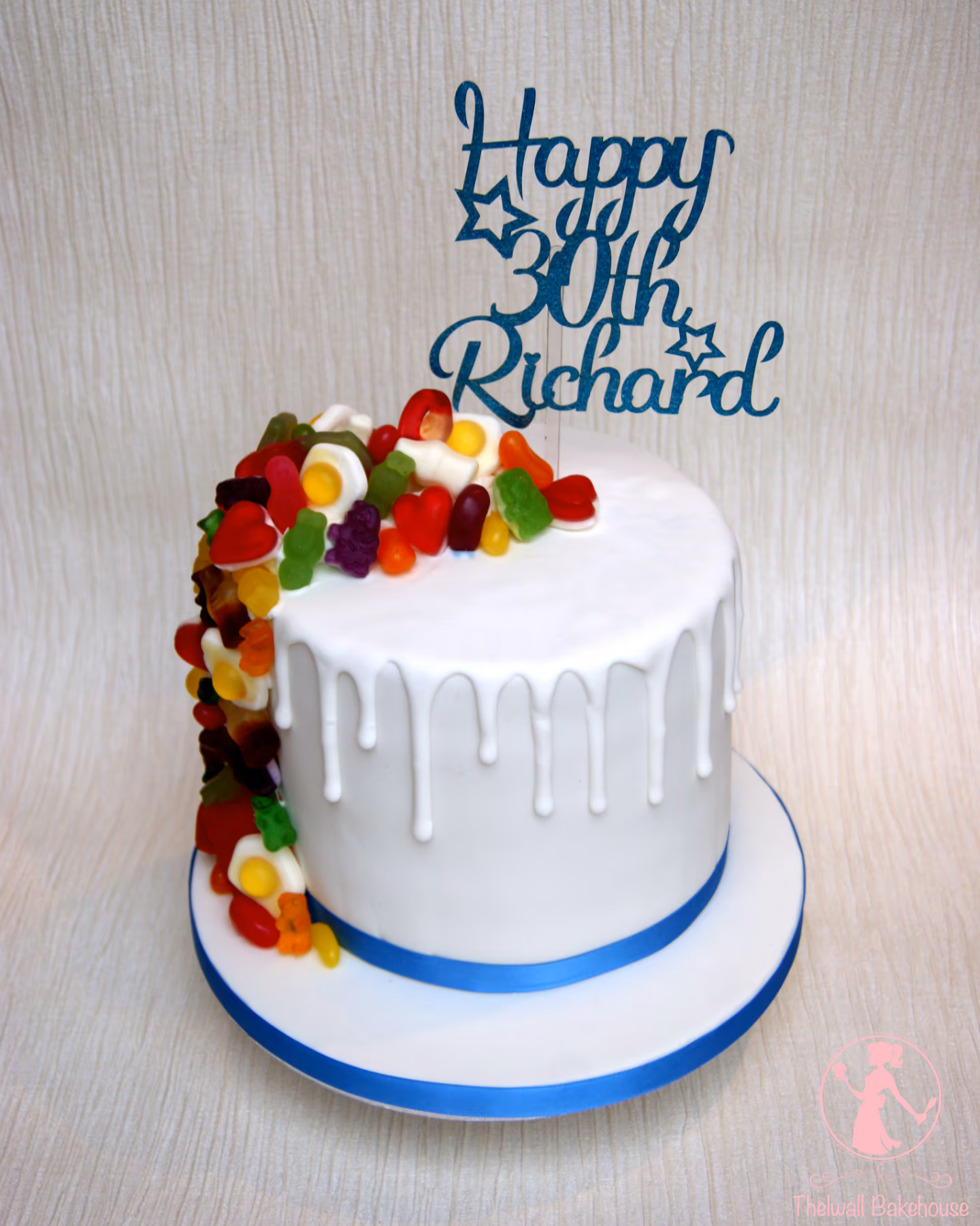 A round white birthday cake with white drip icing and a cascade of colorful gummy candies on one side. A blue glittery cake topper reads "Happy 30th Richard" with star accents. The cake sits on a white board with a blue ribbon.