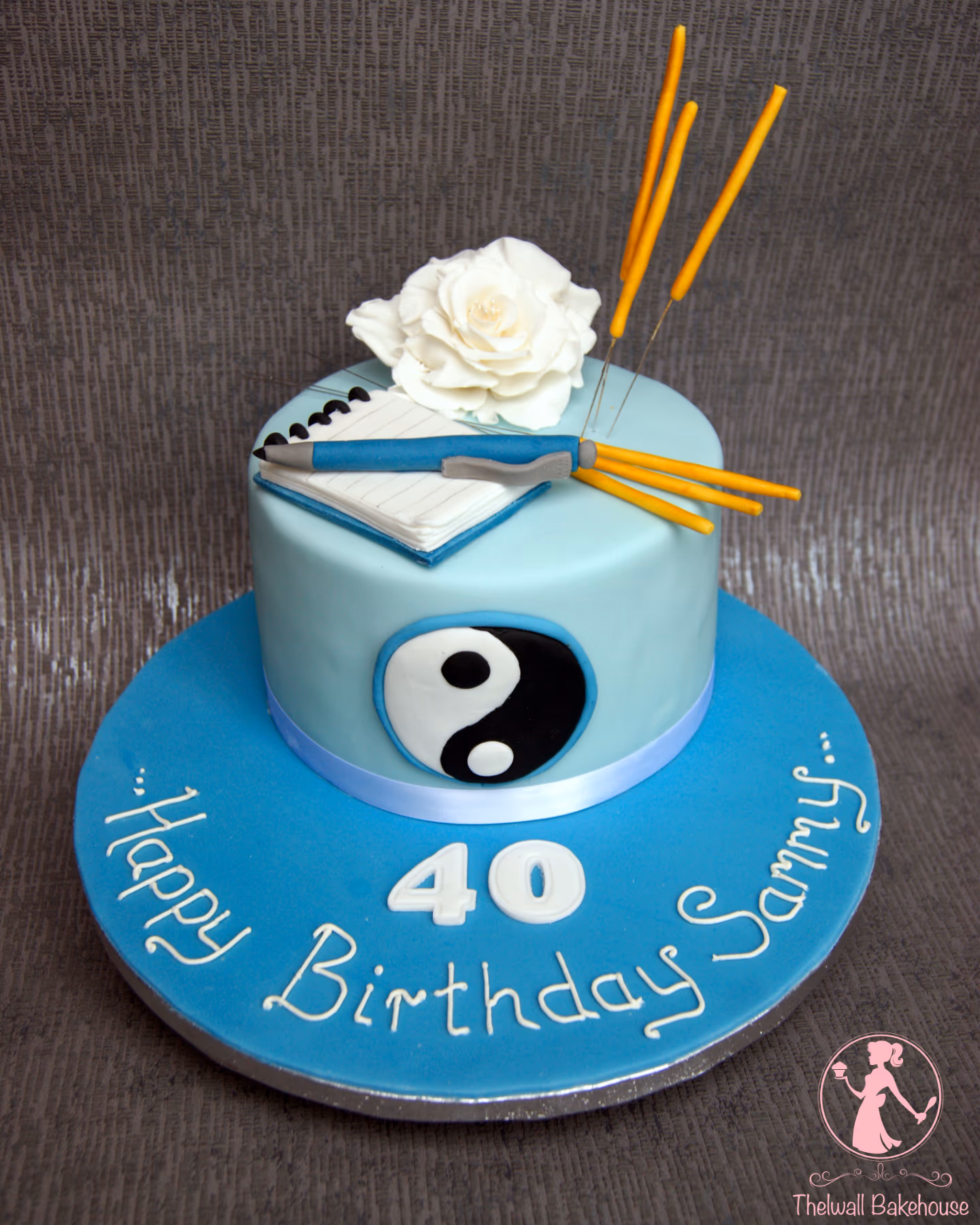 A two-tiered round birthday cake, featuring a light blue top tier and a darker blue base. The top is decorated with a white sugar rose, a white notepad with a blue pen, and several yellow stick decorations. The side of the top tier has a black and white yin-yang symbol, and the blue base is inscribed with 'Happy Birthday Sammy' and the number '40' in white icing.